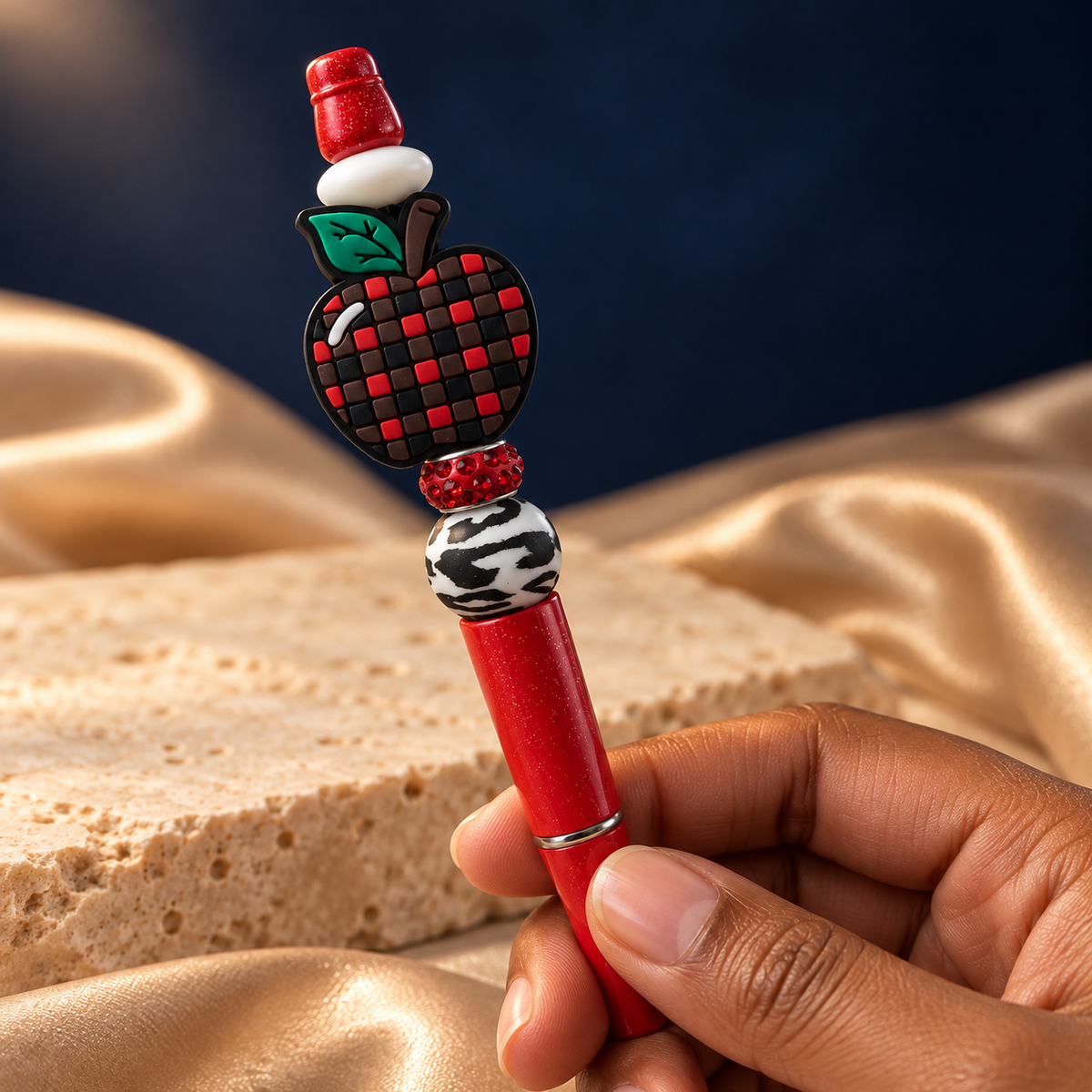 Red glitter-look apple beaded pen held in hand, showing its checkered apple focal piece, green leaf, and black-and-white patterned bead