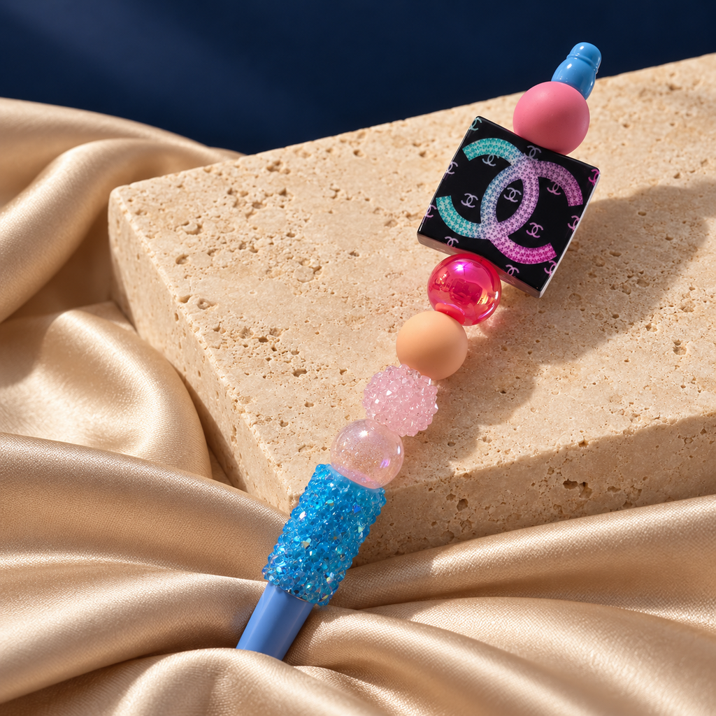 Candy Pop blue pen resting on stone, with pink and peach beads, sparkling blue accents, and colorful double-C charm