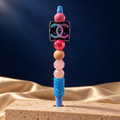 Blue pen with blue rhinestone grip, pink and peach beads, and black square charm featuring colorful interlocking C logos