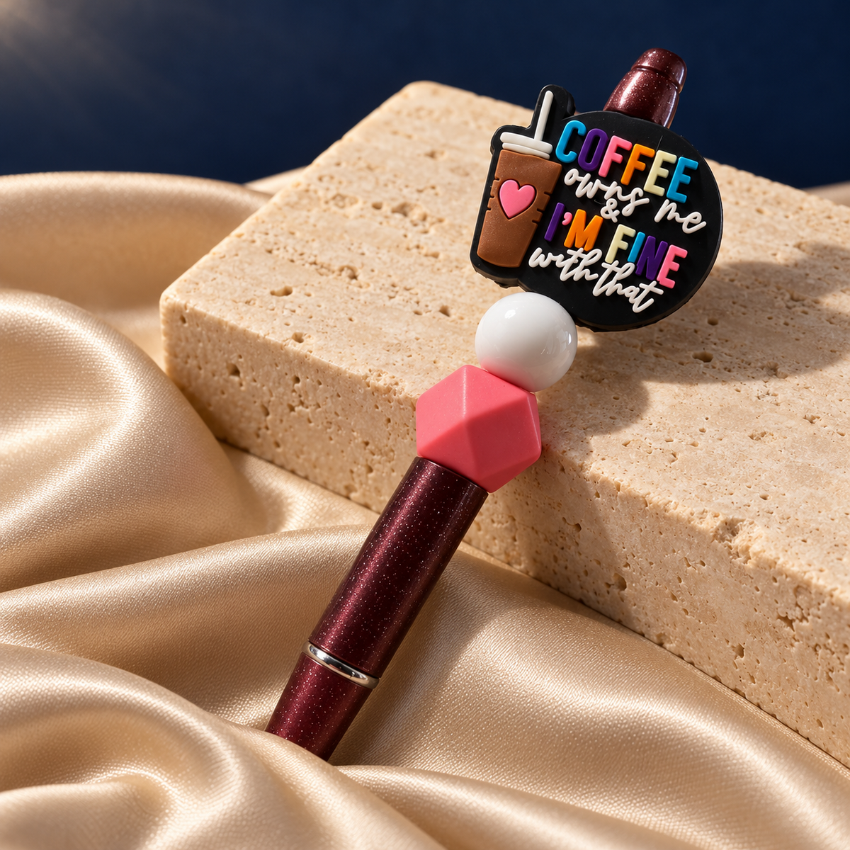 Coffee Mood burgundy glitter-look pen resting on stone, showing a colorful coffee quote charm and pink and white beads