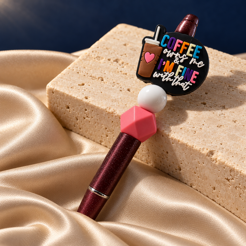 Coffee Mood burgundy glitter-look pen resting on stone, showing a colorful coffee quote charm and pink and white beads