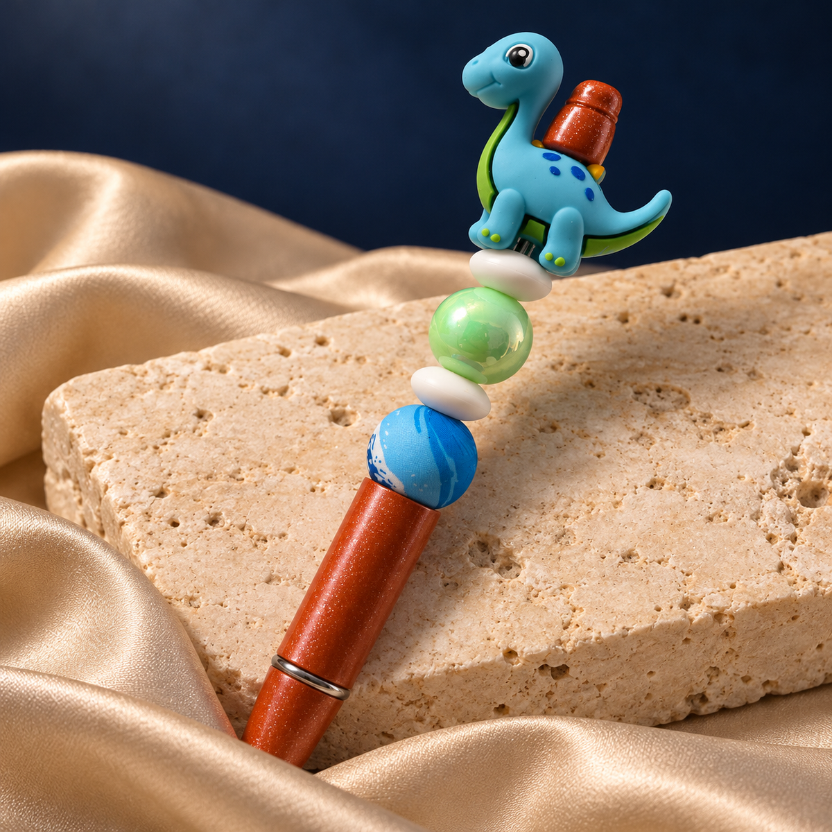 Blue dinosaur beaded pen with orange glitter-look barrel and green, blue, and white beaded accents, resting on a stone display