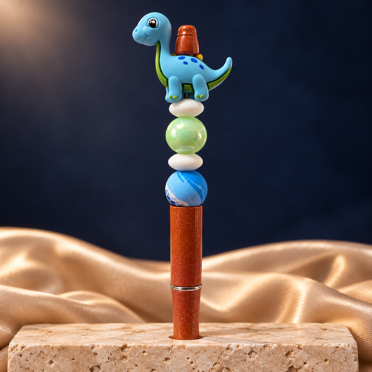 Blue dinosaur beaded pen with an orange glitter-look barrel, green and blue beads, and white spacers, shown upright