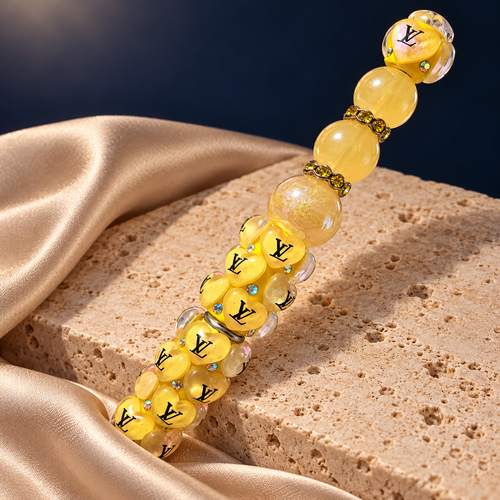 Angled detail of a yellow heart-covered pen with black lettering, translucent beads, and golden sparkling spacers, resting on stone
